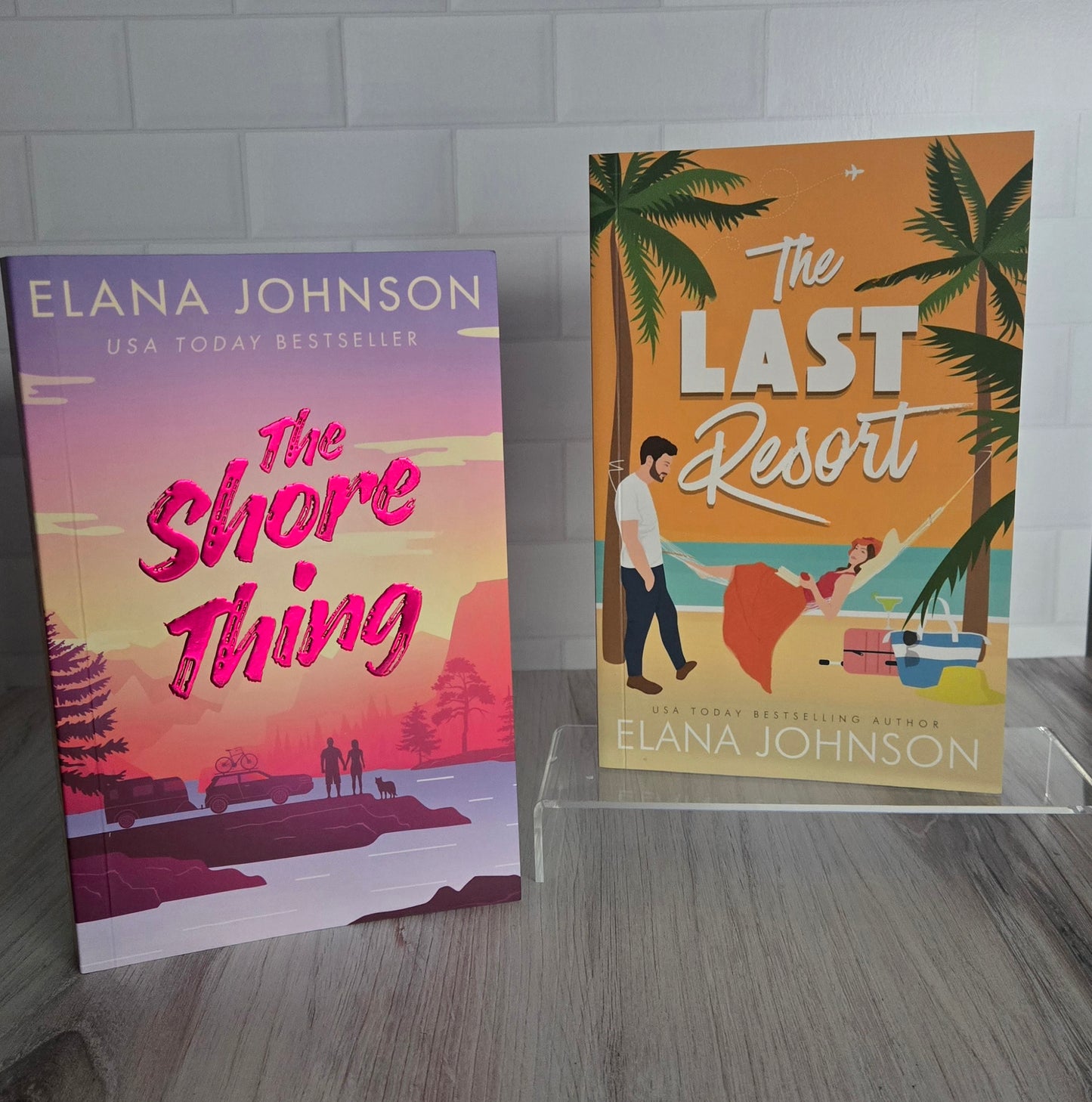 Two books by Elana Johnson on a gray surface with a tiled wall background