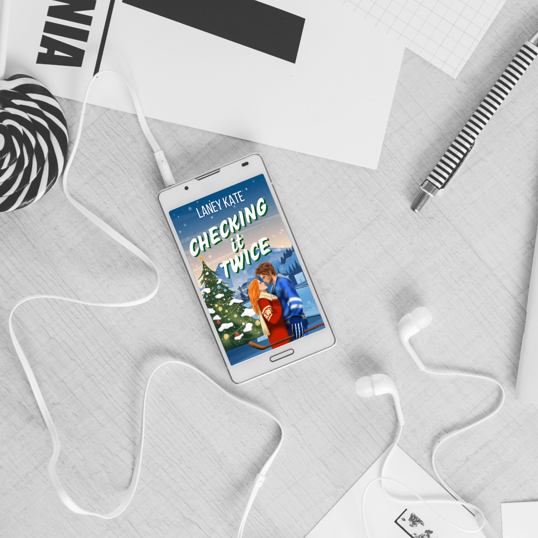 Phone displaying a book cover titled 'Checking It Twice' on a white surface with stationery items.