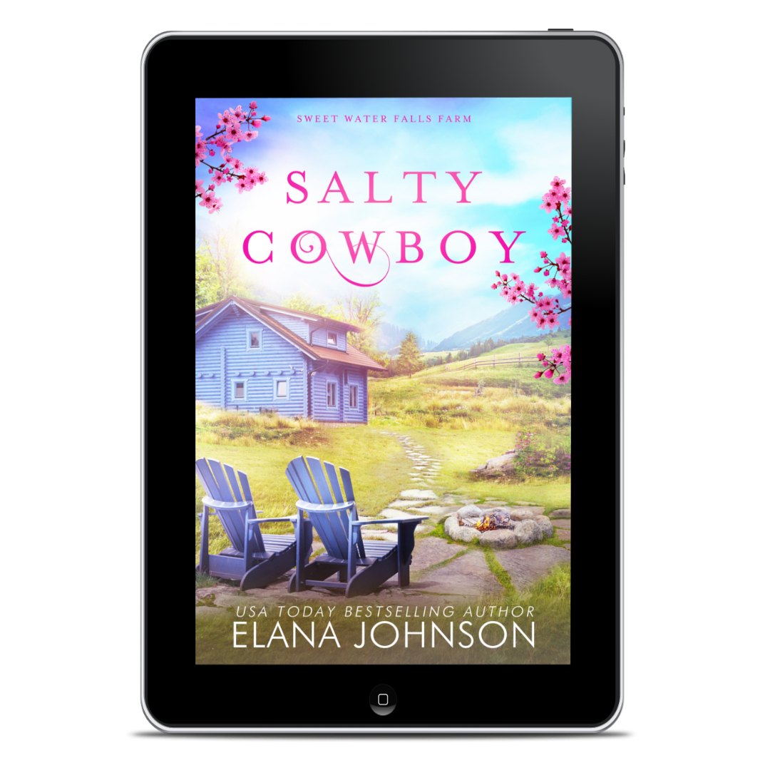 A tablet device centered on a white background, displaying the book cover for "Salty Cowboy" by Elana Johnson. The cover art shows a small blue farmhouse, pink blossoms, two blue Adirondack chairs, a stone fire pit, and a grassy field under a blue sky.