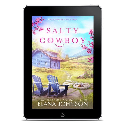 A tablet device centered on a white background, displaying the book cover for "Salty Cowboy" by Elana Johnson. The cover art shows a small blue farmhouse, pink blossoms, two blue Adirondack chairs, a stone fire pit, and a grassy field under a blue sky.